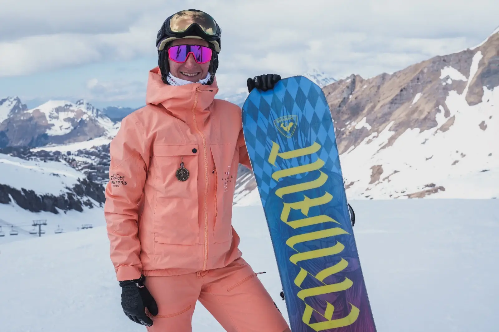 Sofia, souriante, en tenue orange, tient son snowboard debout devant les montagnes.