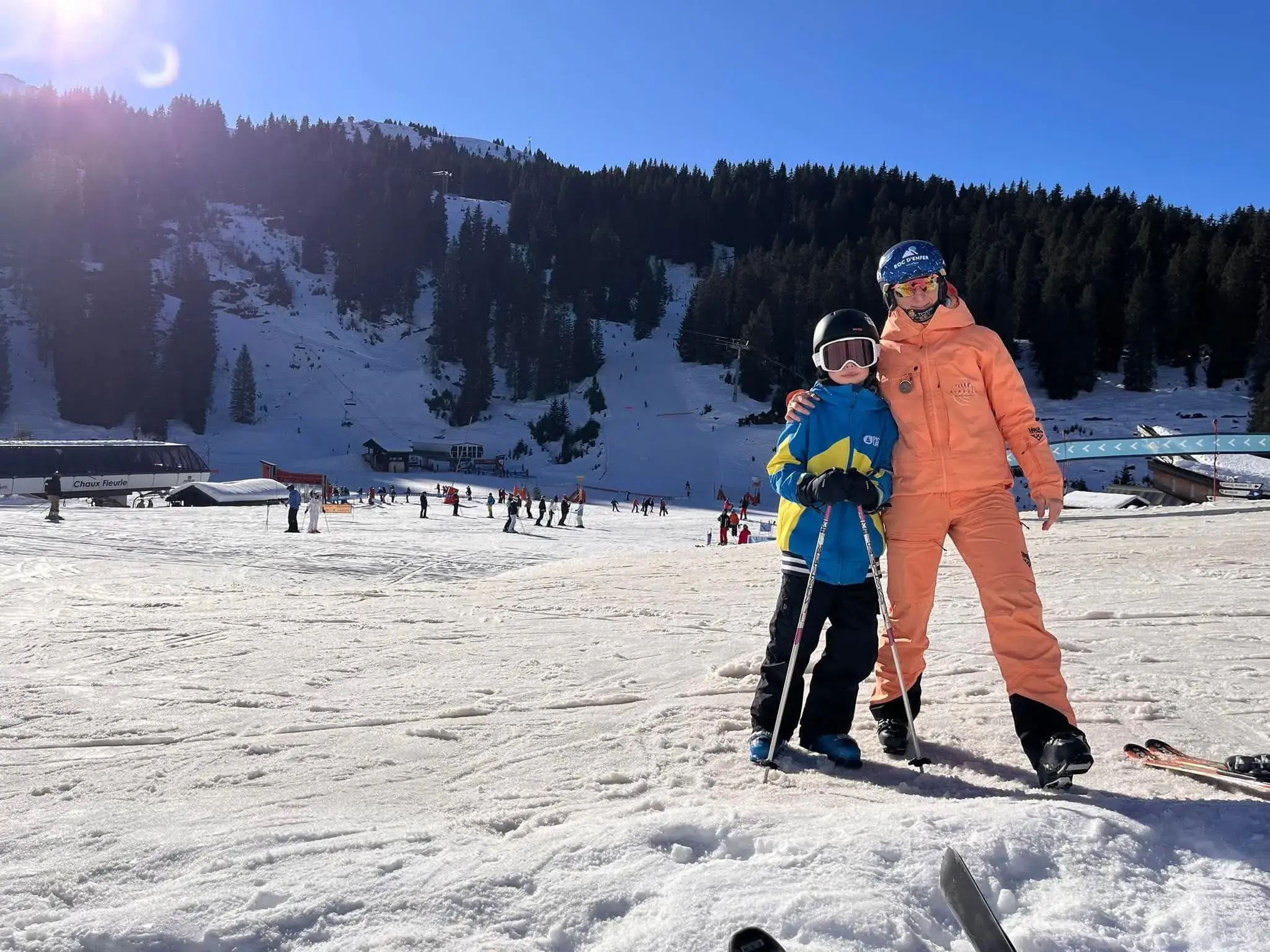 Sofia et un jeune élève, bâtons en main, au pied des pistes sous un ciel bleu.